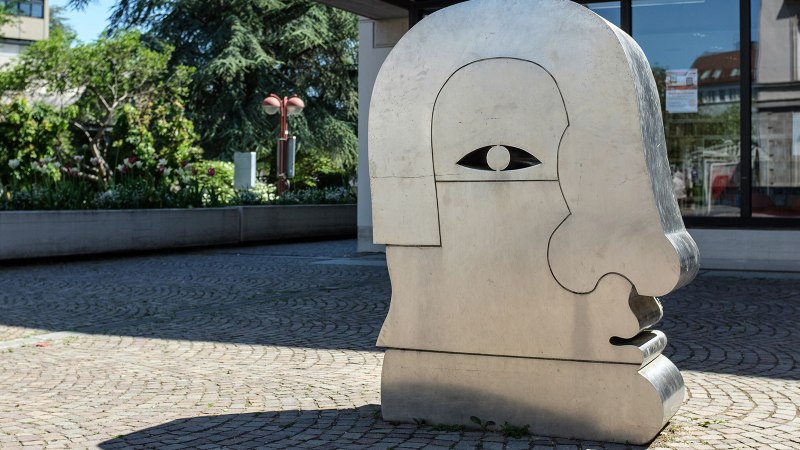 Eine gro&szlig;e Metallskulptur in Form eines stilisierten Kopfes steht auf einem gepflasterten Platz, umgeben von Gr&uuml;npflanzen., &copy; Fotoknobi