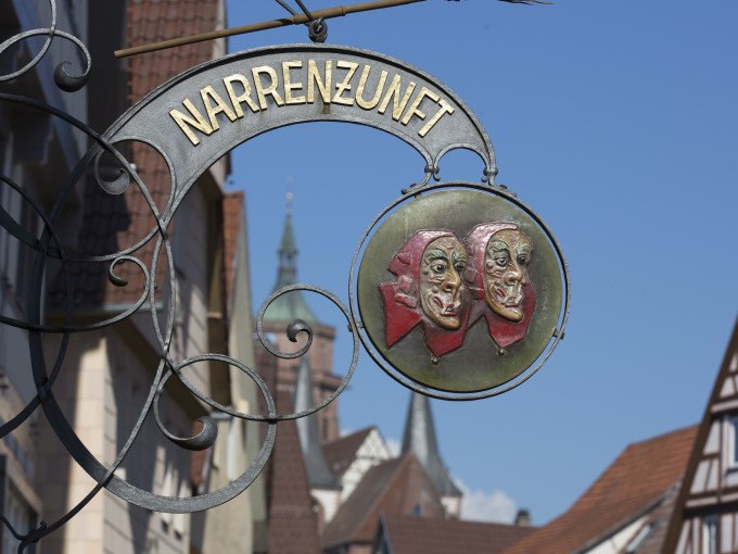 Ein kunstvoll gestaltetes Schild mit der Aufschrift 'NARRNZUNFT' und zwei Masken, im Hintergrund sind Fachwerkh&auml;user und ein Kirchturm zu sehen., &copy; Stadtverwaltung Weil der Stadt
