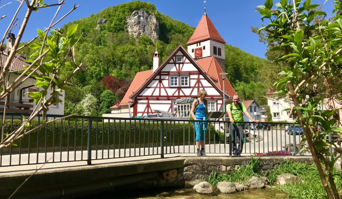Zwei Personen auf einer Brücke vor einem Fachwerkhaus mit Turm. Im Hintergrund grüne Hügel und blauer Himmel. Pflanzen und ein Bach im Vordergrund., © Landkreis Göppingen Zwei Personen auf einer Brücke vor einem Fachwerkhaus mit Turm. Im Hintergrund grüne Hügel und blauer Himmel. Pflanzen und ein Bach im Vordergrund., © Landkreis Göppingen