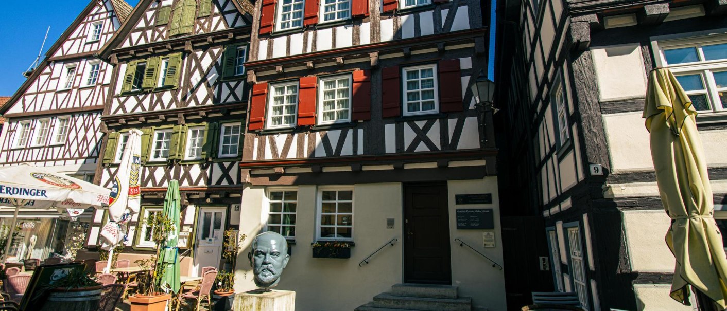 Fachwerkhäuser in Schorndorf mit einer Büste von Gottlieb Daimler im Vordergrund. Sonnenschirme und ein Cafébereich sind ebenfalls zu sehen., © Stuttgart-Marketing GmbH, Sarah Schmid