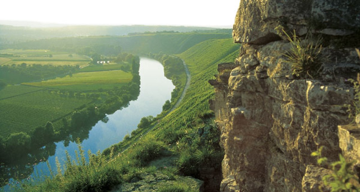 Ein Fluss schlängelt sich durch eine grüne Landschaft, umgeben von Feldern und Hügeln. Im Vordergrund sind Felsen zu sehen., © Land der 1000 Hügel - Kraichgau-Stromberg Ein Fluss schlängelt sich durch eine grüne Landschaft, umgeben von Feldern und Hügeln. Im Vordergrund sind Felsen zu sehen., © Land der 1000 Hügel - Kraichgau-Stromberg