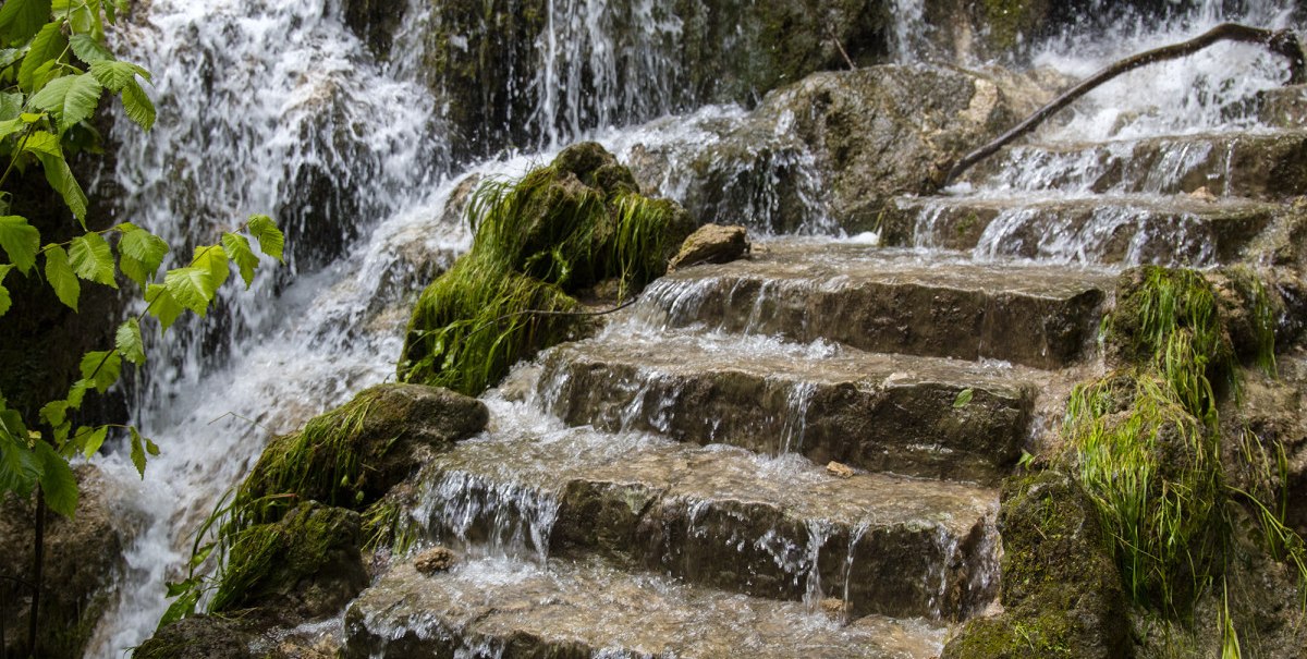 Eine steinerne Treppe mit fließendem Wasser und Moos, umgeben von üppigem Grün am Uracher Wasserfall., © Bad Urach Tourismus