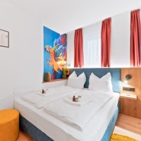 Modernes Doppelzimmer mit weißem Bett, Papageien-Wandbild, roten Vorhängen und gelbem Hocker. Pflegeprodukte auf dem Bett., © JUNGLE N°5 GmbH