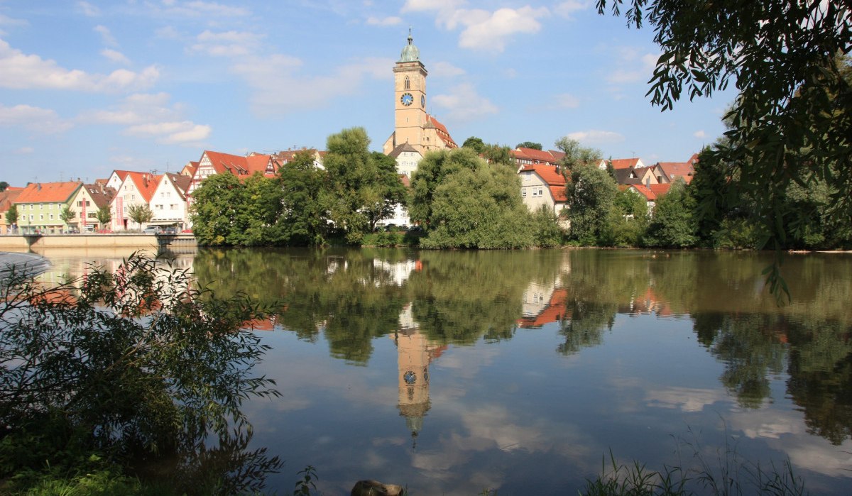 Panorama von Nürtingen mit Kirche und Fachwerkhäusern, die sich im ruhigen Wasser spiegeln, umgeben von Bäumen und blauem Himmel., © Stadt Nürtingen Panorama von Nürtingen mit Kirche und Fachwerkhäusern, die sich im ruhigen Wasser spiegeln, umgeben von Bäumen und blauem Himmel., © Stadt Nürtingen
