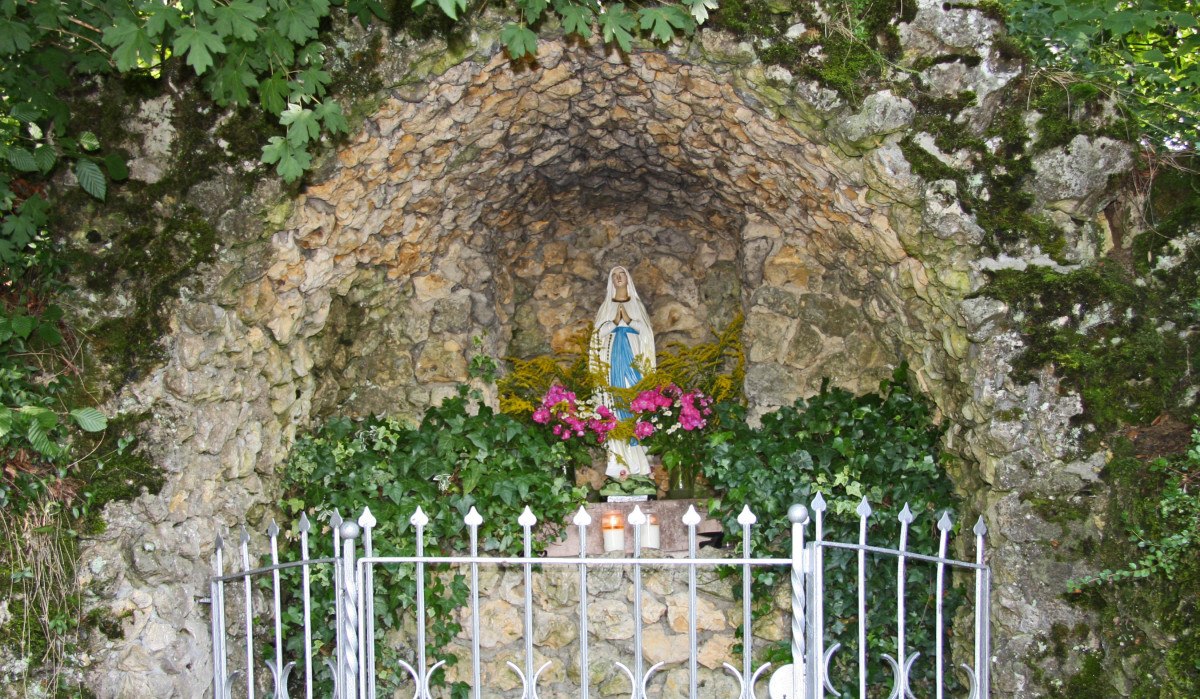 Eine Steingrotte mit einer Marienstatue, umgeben von Blumen und Pflanzen. Ein Metallzaun schützt die Grotte., © Foto: Hans-Werner Kalb