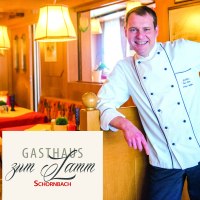 Ein Koch in weißer Uniform steht lächelnd in einem gemütlichen Gasthaus mit Holzmöbeln und roten Tischdecken., © Gasthaus Lamm Schornbach, Fotografie Nicole Schielberg