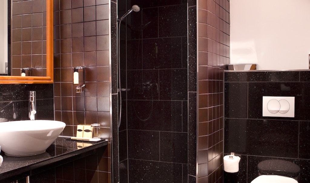 Modernes Badezimmer mit dunklen Fliesen, Waschbecken, Spiegel, Dusche und Toilette., © Ross Messehotel Modernes Badezimmer mit dunklen Fliesen, Waschbecken, Spiegel, Dusche und Toilette., © Ross Messehotel