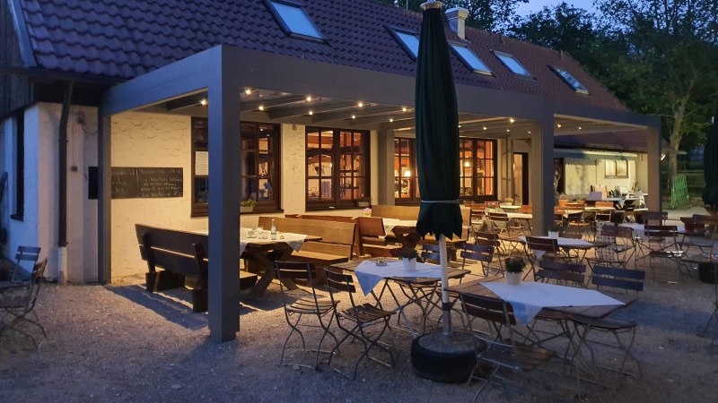 Gem&uuml;tlicher Au&szlig;enbereich eines Restaurants bei D&auml;mmerung, mit Holztischen und -st&uuml;hlen unter einem &uuml;berdachten Bereich. Beleuchtung sorgt f&uuml;r eine warme Atmosph&auml;re., &copy; Kr&auml;uterb&uuml;hl