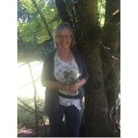 Eine Frau steht l&auml;chelnd neben einem Baum und h&auml;lt einen Strau&szlig; Wildkr&auml;uter. Sonnenlicht f&auml;llt durch die Bl&auml;tter und erzeugt ein angenehmes Lichtspiel., &copy; Touristik und Marketing GmbH