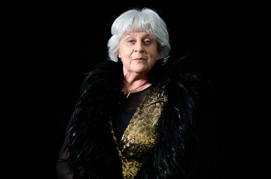 Ältere Frau mit weißem Haar, goldener Kleidung und schwarzem Federumhang vor dunklem Hintergrund.