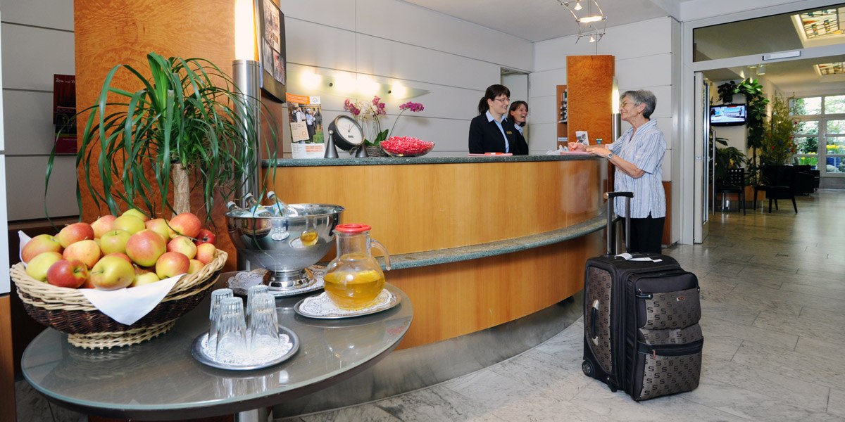 Eine Hotelrezeption mit zwei Mitarbeiterinnen, die einer älteren Frau mit Koffer helfen. Ein Tisch mit Äpfeln und Getränken steht im Vordergrund., © Hotel Unger
