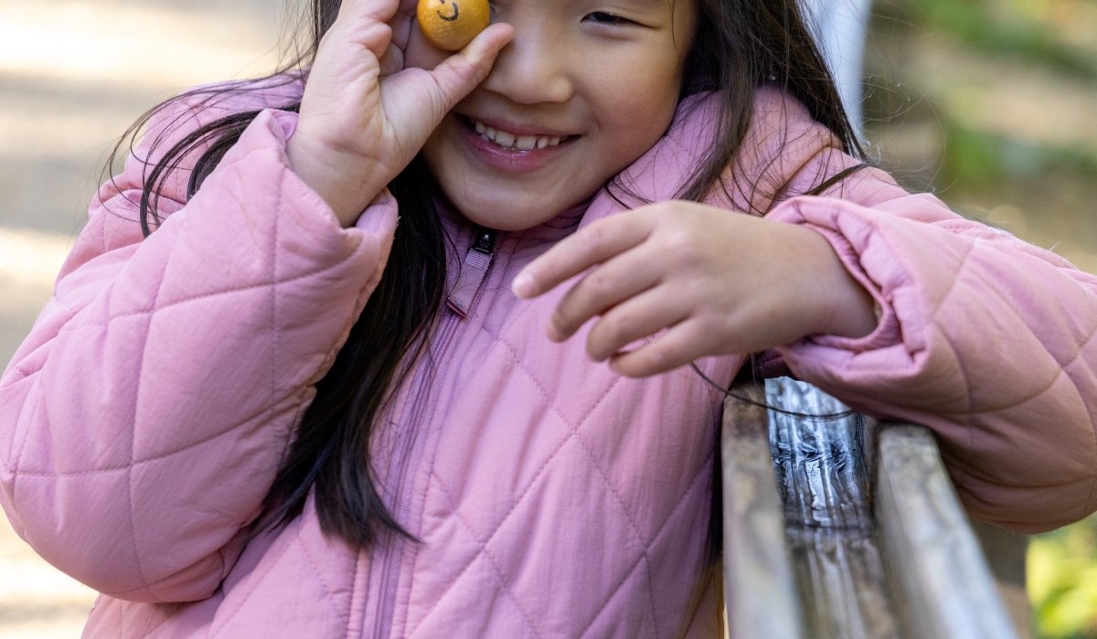 Ein Kind in rosa Jacke hält eine kleine Kugel mit Gesicht vor das Auge und lächelt fröhlich. Im Hintergrund ist ein Weg zu sehen., © Thomas Zehnder
