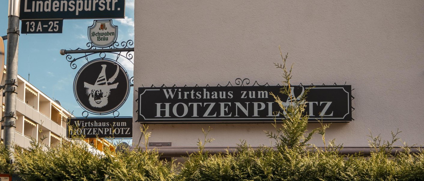Schild des Wirtshauses zum Hotzenplotz an der Lindenspürstraße, umgeben von grünen Büschen und einem blauen Himmel im Hintergrund., © SMG. Sarah Schmid Schild des Wirtshauses zum Hotzenplotz an der Lindenspürstraße, umgeben von grünen Büschen und einem blauen Himmel im Hintergrund., © SMG. Sarah Schmid