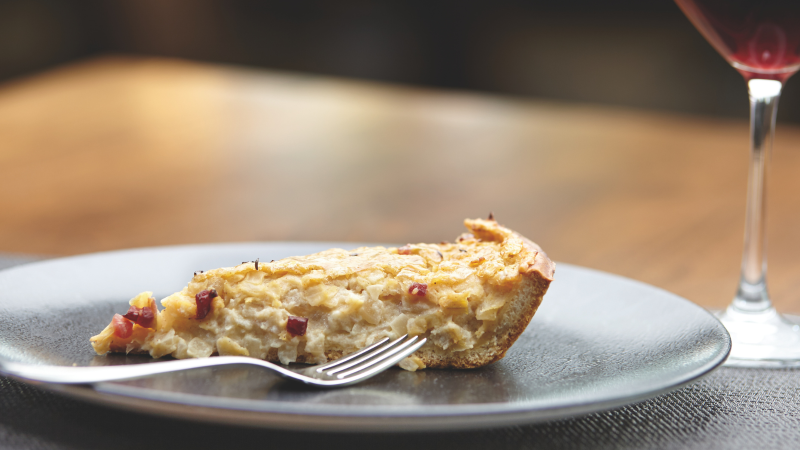 Ein Stück Zwiebelkuchen auf einem Teller mit einer Gabel, daneben ein Glas Rotwein auf einem Holztisch., © SMG Jean-Claude Winkler
