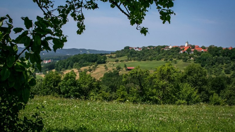 Grüne Wiesen und Bäume rahmen ein Dorf mit roten Dächern und einer Kirche im Hintergrund ein. Der Himmel ist klar und blau., © agentur arcos/Niki Eilers Grüne Wiesen und Bäume rahmen ein Dorf mit roten Dächern und einer Kirche im Hintergrund ein. Der Himmel ist klar und blau., © agentur arcos/Niki Eilers
