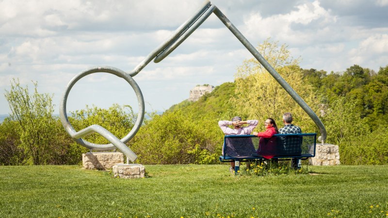 Drei Personen sitzen auf einer Bank vor einer großen Brillenskulptur in einer grünen Landschaft mit Hügeln und Wolken am Himmel., © hochgehberge