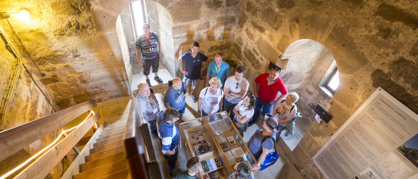 Eine Gruppe von Menschen steht in einem historischen Turm mit Steinwänden und betrachtet eine Ausstellung. Es gibt Treppen und Informationstafeln., © 8pm.de