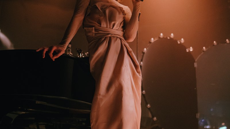 Eine Frau in einem eleganten Kleid lehnt an einem Klavier und singt in ein Mikrofon, umgeben von warmem Bühnenlicht., © Ann-Marie Schwanke Eine Frau in einem eleganten Kleid lehnt an einem Klavier und singt in ein Mikrofon, umgeben von warmem Bühnenlicht., © Ann-Marie Schwanke