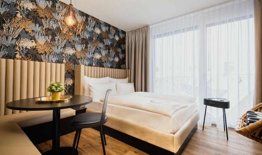 Modernes Apartmentzimmer mit stilvoller Tapete, gem&uuml;tlichem Bett, rundem Tisch und Stuhl. Gro&szlig;e Fenster mit Vorh&auml;ngen lassen viel Licht herein., &copy; Rioca Stuttgart Posto 4 GmbH