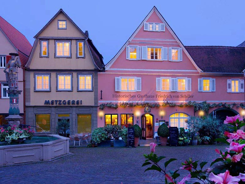 Ein malerisches Dorf bei Dämmerung mit einem historischen Gasthaus und einer Metzgerei. Blumen und ein Brunnen schmücken den Platz. Ein malerisches Dorf bei Dämmerung mit einem historischen Gasthaus und einer Metzgerei. Blumen und ein Brunnen schmücken den Platz.