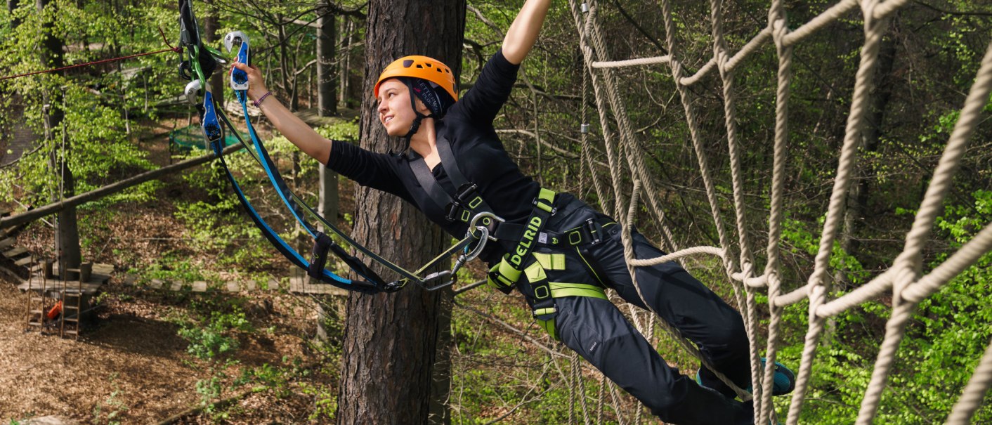 Eine Person in einem Hochseilgarten, gesichert mit Helm und Klettergurt, balanciert auf einem Seil zwischen Bäumen., © Kletterwald Plochingen Eine Person in einem Hochseilgarten, gesichert mit Helm und Klettergurt, balanciert auf einem Seil zwischen Bäumen., © Kletterwald Plochingen