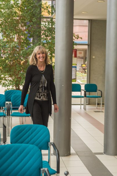 Eine Frau in schwarzer Kleidung geht lächelnd durch einen Raum mit blauen Stühlen und Pflanzen im Hintergrund.