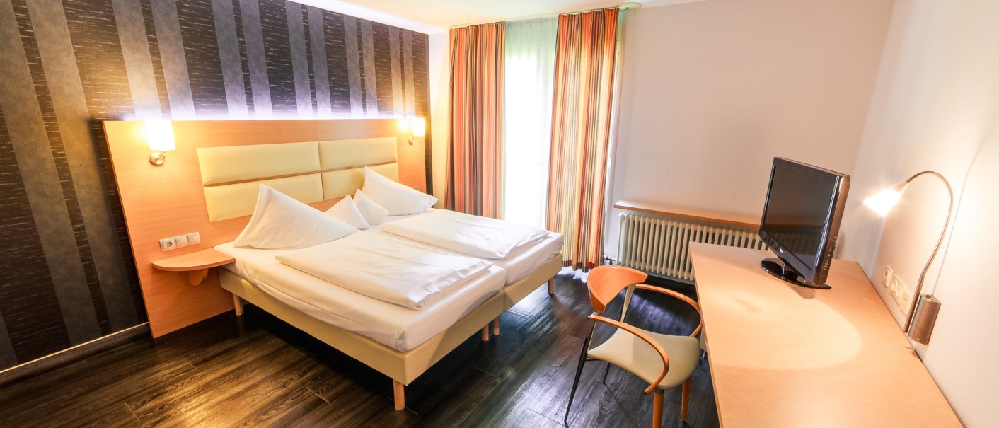 Modernes Doppelzimmer mit großem Bett, Schreibtisch, Stuhl und Fernseher. Die Wände sind teils dunkel gestreift, Vorhänge in warmen Farben., © PLAZA Hotelgroup GmbH Modernes Doppelzimmer mit großem Bett, Schreibtisch, Stuhl und Fernseher. Die Wände sind teils dunkel gestreift, Vorhänge in warmen Farben., © PLAZA Hotelgroup GmbH