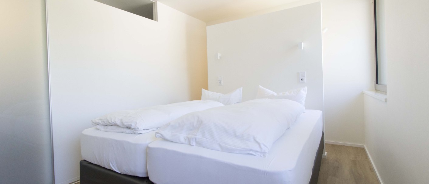Helles Schlafzimmer mit einem großen Doppelbett, weißen Bettbezügen, Holzboden und einem Fenster an der rechten Wand., © Büroma-Apart Apartmentvermietung GmbH Helles Schlafzimmer mit einem großen Doppelbett, weißen Bettbezügen, Holzboden und einem Fenster an der rechten Wand., © Büroma-Apart Apartmentvermietung GmbH
