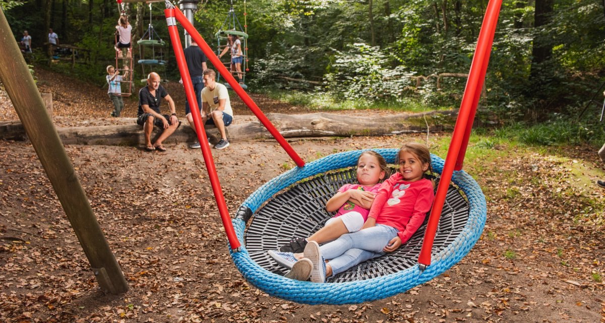 Zwei Kinder entspannen auf einer großen Netzschaukel im Waldspielplatz. Im Hintergrund spielen weitere Kinder, während Erwachsene auf einem Baumstamm sitzen. Zwei Kinder entspannen auf einer großen Netzschaukel im Waldspielplatz. Im Hintergrund spielen weitere Kinder, während Erwachsene auf einem Baumstamm sitzen.