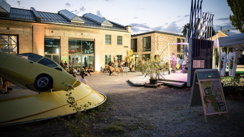 Abendveranstaltung im Kunstverein Wagenhalle mit Kunstinstallation eines gelben Autos und Menschen, die auf dem Platz sitzen und einer Darbietung zuschauen., © Atelier Oradoro Abendveranstaltung im Kunstverein Wagenhalle mit Kunstinstallation eines gelben Autos und Menschen, die auf dem Platz sitzen und einer Darbietung zuschauen., © Atelier Oradoro