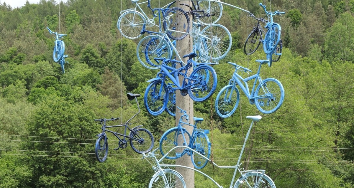 Blau gestrichene Fahrräder hängen als Kunstinstallation an einem Pfosten in einer grünen, bewaldeten Landschaft., © Natur.Nah. Schönbuch & Heckengäu Blau gestrichene Fahrräder hängen als Kunstinstallation an einem Pfosten in einer grünen, bewaldeten Landschaft., © Natur.Nah. Schönbuch & Heckengäu