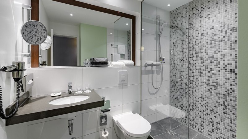 Modernes Badezimmer mit großer Dusche, Waschbecken, Spiegel und Toilette. Die Wände sind teils mit Mosaikfliesen verkleidet., © Mercure Hotel Stuttgart Sindelfingen an der Messe