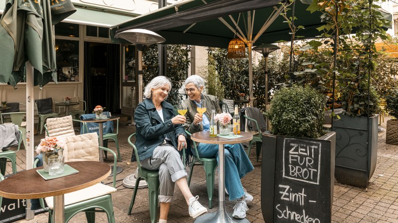 Zwei Frauen sitzen in einem gemütlichen Café im Freien, stoßen mit Getränken an und lachen. Ein Schild bietet Zimtschnecken an., © Stuttgart Marketing GmbH - Sarah Schmid Zwei Frauen sitzen in einem gemütlichen Café im Freien, stoßen mit Getränken an und lachen. Ein Schild bietet Zimtschnecken an., © Stuttgart Marketing GmbH - Sarah Schmid