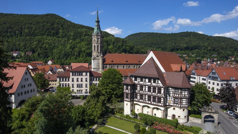 Stadtansicht mit Fachwerkh&auml;usern und Kirchturm, umgeben von gr&uuml;nen H&uuml;geln und blauem Himmel., &copy; Bad Urach Tourismus