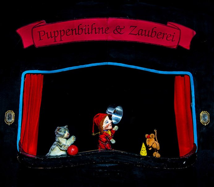 Eine Puppenbühne zeigt eine Katze, eine Puppe mit einem Topf und eine Maus mit Käse. Der Vorhang ist rot, der Hintergrund schwarz., © Prinzessin Gisela Theater