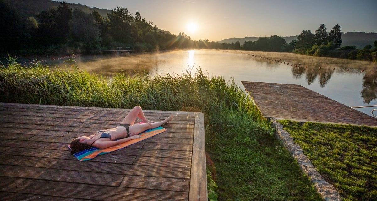 Eine Person liegt auf einem Steg am See und genießt den Sonnenaufgang. Der See ist von Bäumen umgeben und es steigt Nebel auf., © Stoppel Eine Person liegt auf einem Steg am See und genießt den Sonnenaufgang. Der See ist von Bäumen umgeben und es steigt Nebel auf., © Stoppel