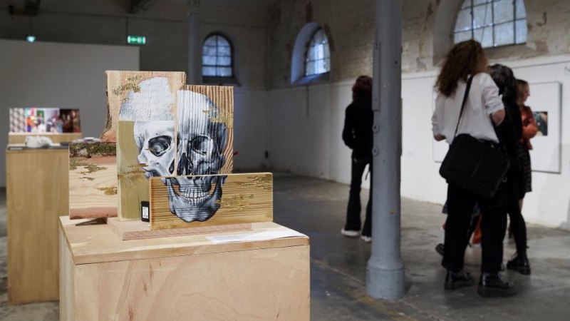 Eine Kunstinstallation mit einem Totenkopf auf Holzbl&ouml;cken in einer Galerie. Im Hintergrund sind Besucher zu sehen, die die Ausstellung betrachten., &copy; Kunstzentrum Karlskaserne