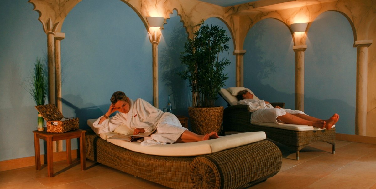 Zwei Personen in weißen Bademänteln entspannen auf Liegen in einem stilvollen Spa-Raum mit blauen Wänden und dekorativen Säulen., © TOMAS