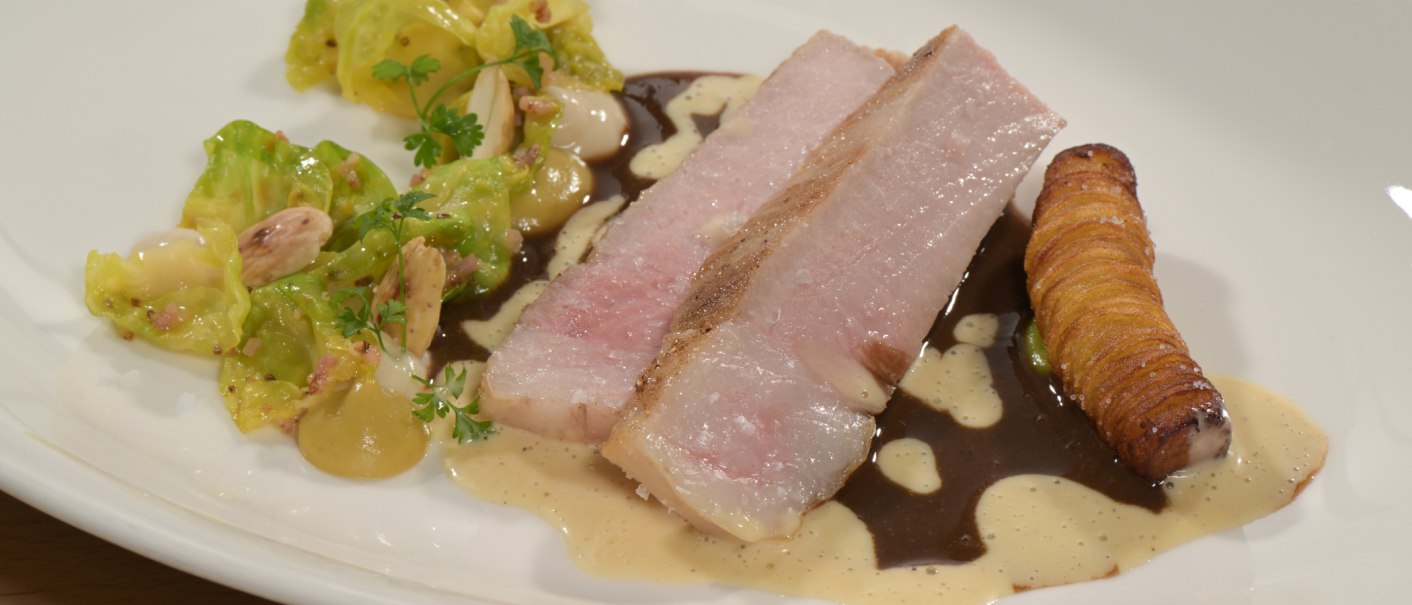 Gourmetgericht mit Schweinefleisch, knuspriger Kartoffelrolle, grünem Gemüse und cremiger Sauce auf einem weißen Teller angerichtet., © Landgasthof + Hotel Jagstmühle Gourmetgericht mit Schweinefleisch, knuspriger Kartoffelrolle, grünem Gemüse und cremiger Sauce auf einem weißen Teller angerichtet., © Landgasthof + Hotel Jagstmühle