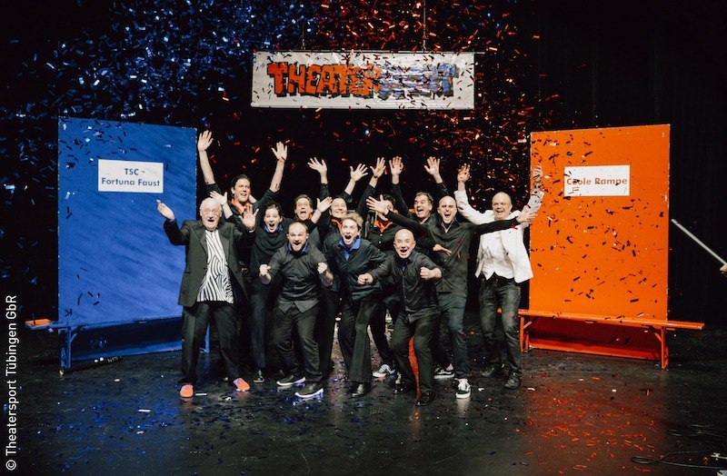 Eine Gruppe von Menschen jubelt auf einer Bühne, umgeben von blauem und rotem Konfetti. Im Hintergrund steht ein Theatersport-Schild., © Stadt Kornwestheim Kulturmanagement Eine Gruppe von Menschen jubelt auf einer Bühne, umgeben von blauem und rotem Konfetti. Im Hintergrund steht ein Theatersport-Schild., © Stadt Kornwestheim Kulturmanagement
