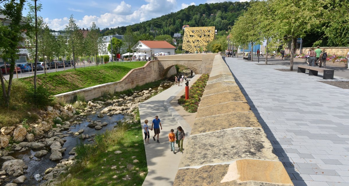 Fußweg entlang eines kleinen Flusses, gesäumt von Bäumen und Blumen. Moderne Architektur im Hintergrund, sonniges Wetter., © Burkhard Walther Fußweg entlang eines kleinen Flusses, gesäumt von Bäumen und Blumen. Moderne Architektur im Hintergrund, sonniges Wetter., © Burkhard Walther