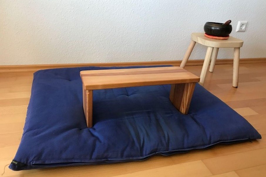Ein Meditationskissen mit einem Holzhocker darauf, daneben ein kleiner Tisch mit einer Klangschale auf einem Holzboden., © Bürgerengagement und Bürgerhäuser