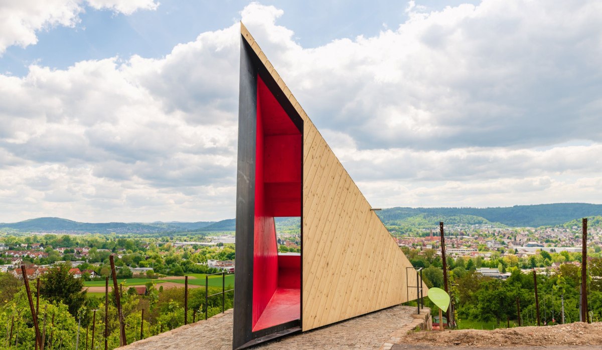 Moderne Holzstruktur mit rotem Innenraum auf einem Hügel. Im Hintergrund eine weite Landschaft mit Hügeln und Wolken am Himmel., © Authentic Studios Moderne Holzstruktur mit rotem Innenraum auf einem Hügel. Im Hintergrund eine weite Landschaft mit Hügeln und Wolken am Himmel., © Authentic Studios