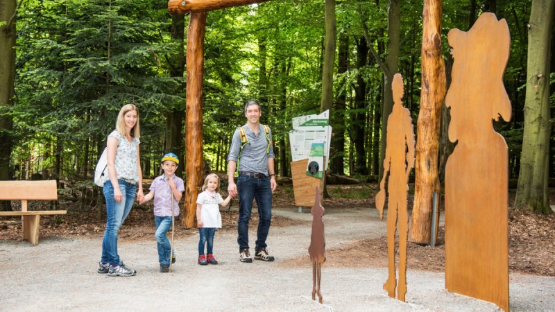 Eine Familie steht am Eingang des Märchenwegs 'Das kalte Herz' in Bad Wildbad, umgeben von Bäumen und Metallfiguren., © Nördlicher Schwarzwald Eine Familie steht am Eingang des Märchenwegs 'Das kalte Herz' in Bad Wildbad, umgeben von Bäumen und Metallfiguren., © Nördlicher Schwarzwald