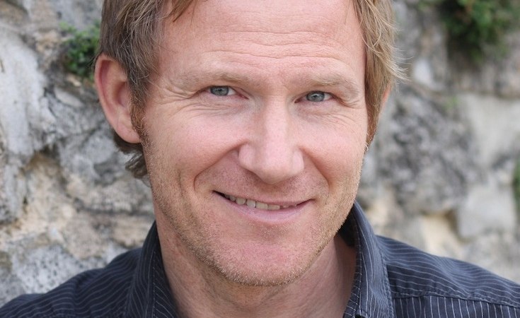 Ein Mann mit kurzen, blonden Haaren lächelt in die Kamera. Er trägt ein dunkles Hemd und steht vor einer Steinmauer mit etwas Grünbewuchs., © Scala Kultur Live gGmbH