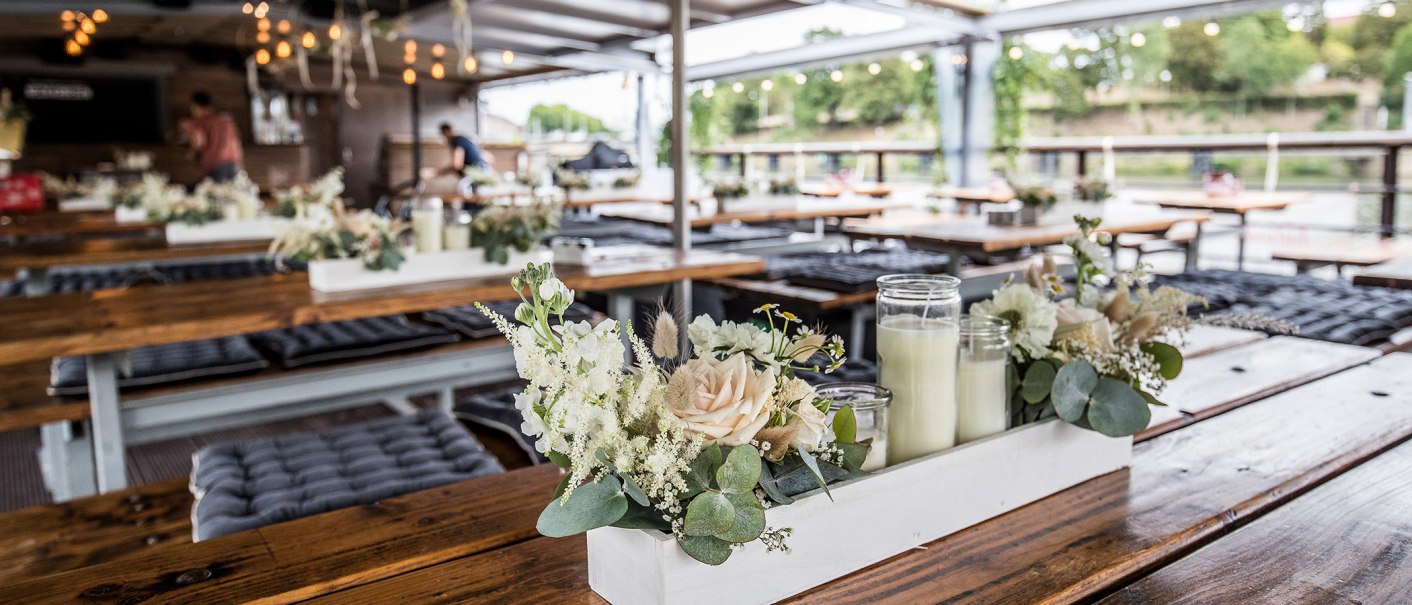 Innenbereich eines Restaurants mit Holztischen, Blumenarrangements und Kerzen. Im Hintergrund sind Lichter und eine Person zu sehen., © all copyrights are reserved by maks richter Innenbereich eines Restaurants mit Holztischen, Blumenarrangements und Kerzen. Im Hintergrund sind Lichter und eine Person zu sehen., © all copyrights are reserved by maks richter