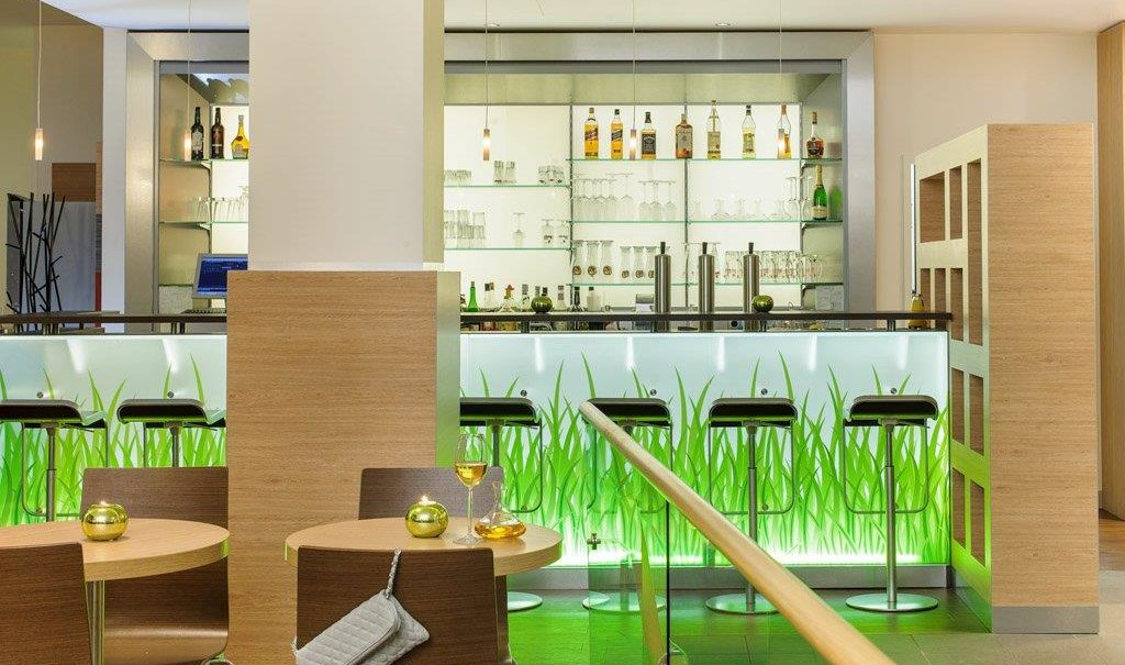 Moderne Hotelbar mit beleuchtetem Tresen, Barhockern und Regalen voller Flaschen. Tische mit Gläsern und Dekoration im Vordergrund., © TOMAS