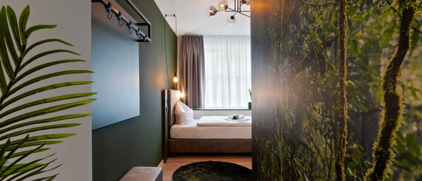 Modernes Einzelzimmer mit grünem Thema, Pflanzenmotiv an der Wand, stilvoller Beleuchtung und gemütlichem Bett., © JUNGLE N°5 GmbH Modernes Einzelzimmer mit grünem Thema, Pflanzenmotiv an der Wand, stilvoller Beleuchtung und gemütlichem Bett., © JUNGLE N°5 GmbH