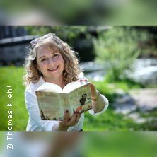 Eine Frau mit lockigem Haar liest ein Buch im Freien und l&auml;chelt in die Kamera. Im Hintergrund ist eine gr&uuml;ne Wiese zu sehen., &copy; links im Bild
