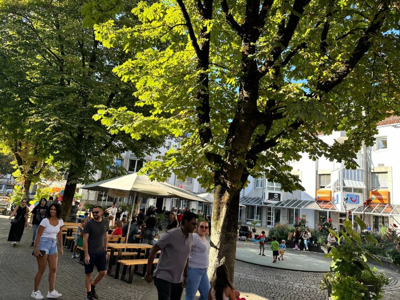 Menschen flanieren an einem sonnigen Tag durch eine belebte Einkaufsstra&szlig;e. B&auml;ume spenden Schatten, w&auml;hrend Gesch&auml;fte und Caf&eacute;s zum Verweilen einladen.